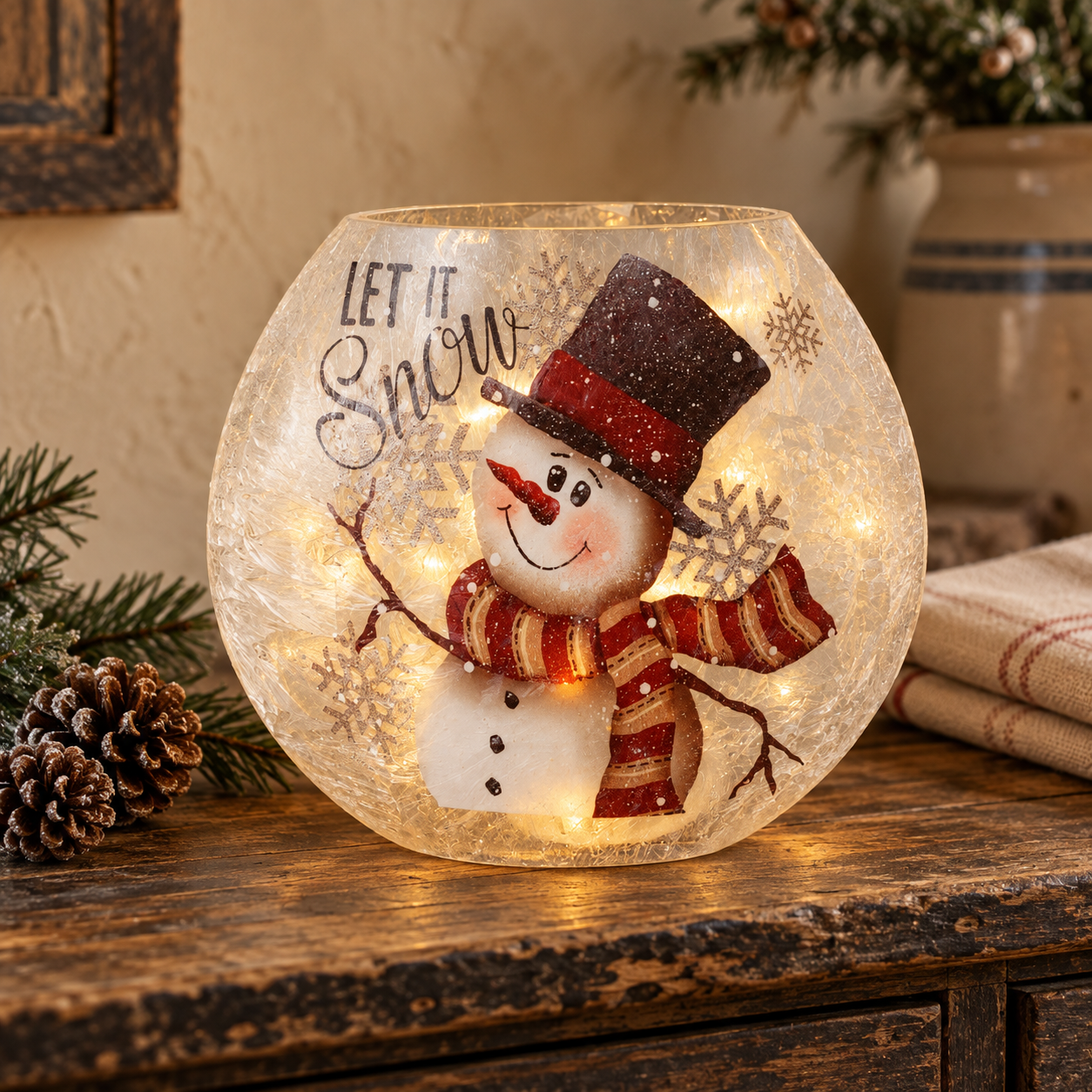 Glowing frosted glass snowman luminary with “Let It Snow” lettering, styled with evergreen sprigs on an aged cupboard.