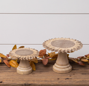 Two small sunflower-shaped pedestal risers with pale carved tops and wooden bases beside autumn leaves.